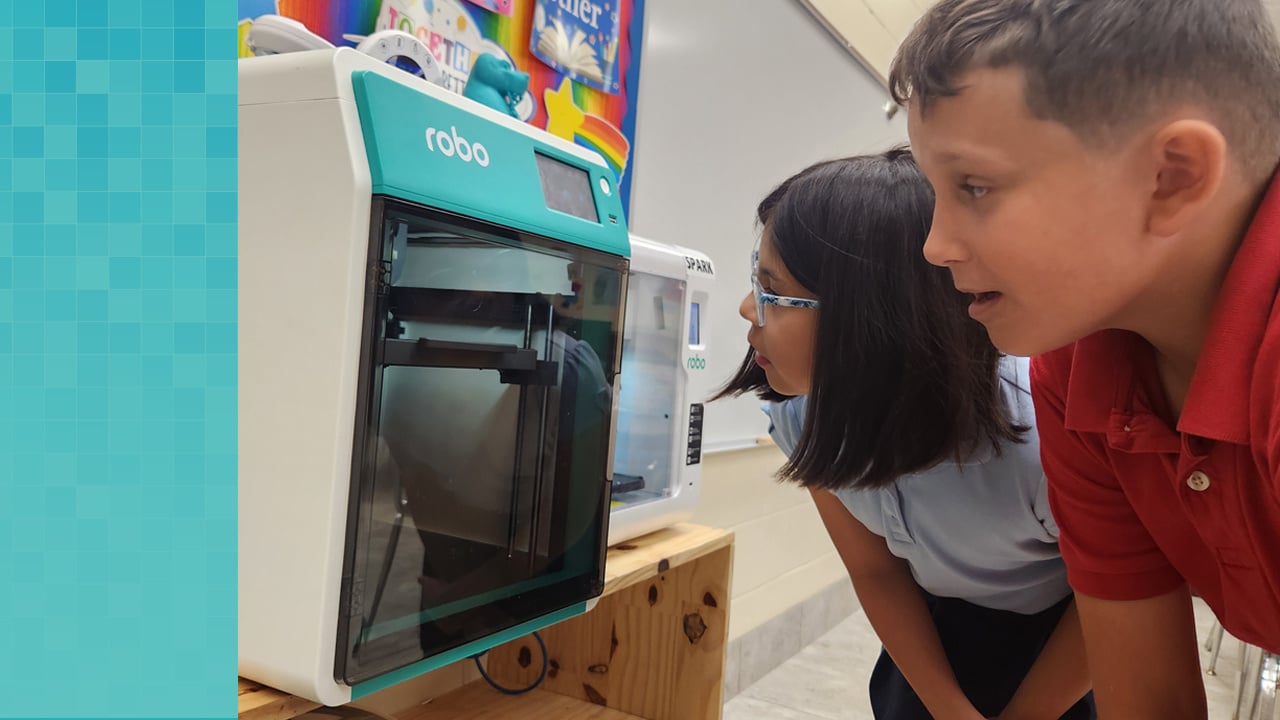 Two elementary-age students look at the printing progress of their design through the Robo E4 3D printer’s glass door.