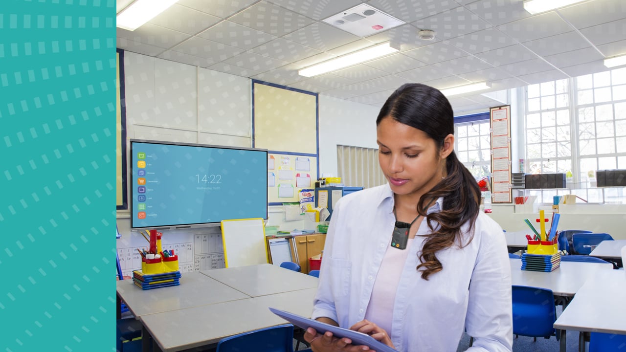 A classroom with a teacher wearing a FrontRow microphone and tapping a tablet screen. An ezRoom speaker is mounted on the ceiling.