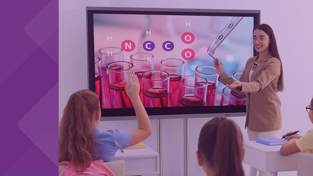 Teacher in front of Boxlight interactive display, pointing to website image showing chemical bonds and glass beakers. A student is raising her hand to participate in the class discussion.