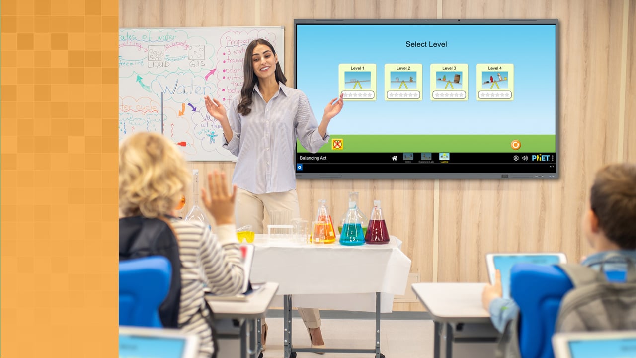 Teacher standing next to Boxlight interactive display showing a game available in MyClass for enhanced student engagement. A student with raised hand is prepared to provide an answer.