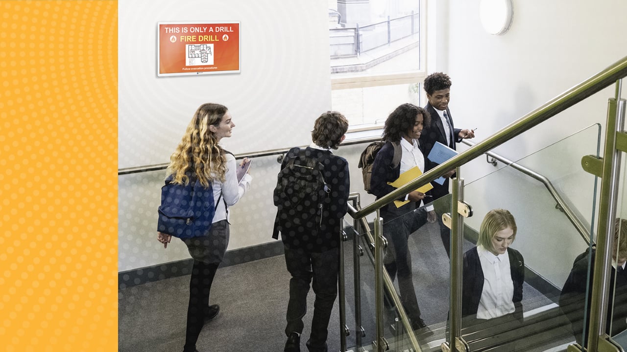 High school students walking through a hallway. TimeSign screen shows Fire Drill alert. 